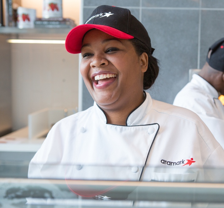 An IN2WORK graduate dressed in a white chef's coat and Aramark hat standing behind a food service station.